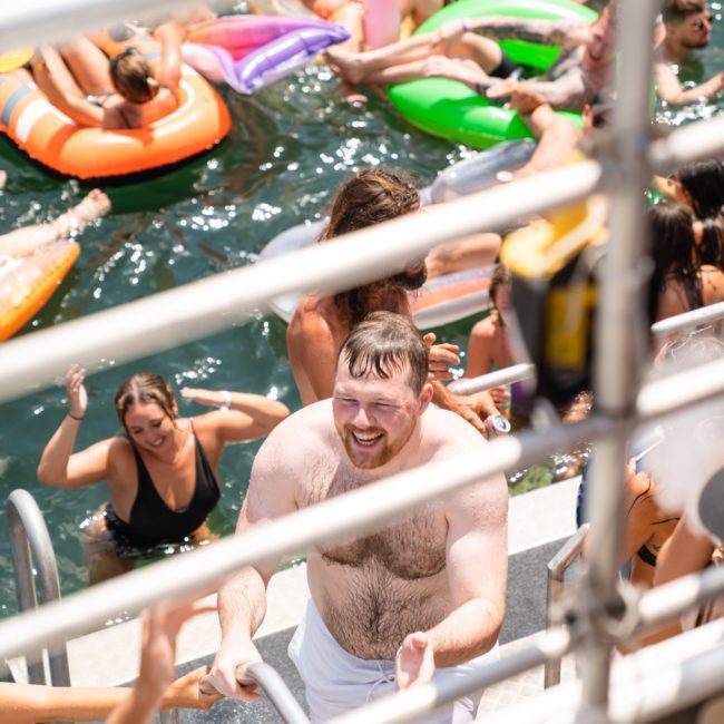 A man climbs a boat ladder from the water as people on inflatable rafts and floaties enjoy a sunny day. Nearby, others indulge in the ultimate relaxation with DJ boat hire Sydney for an unforgettable experience.