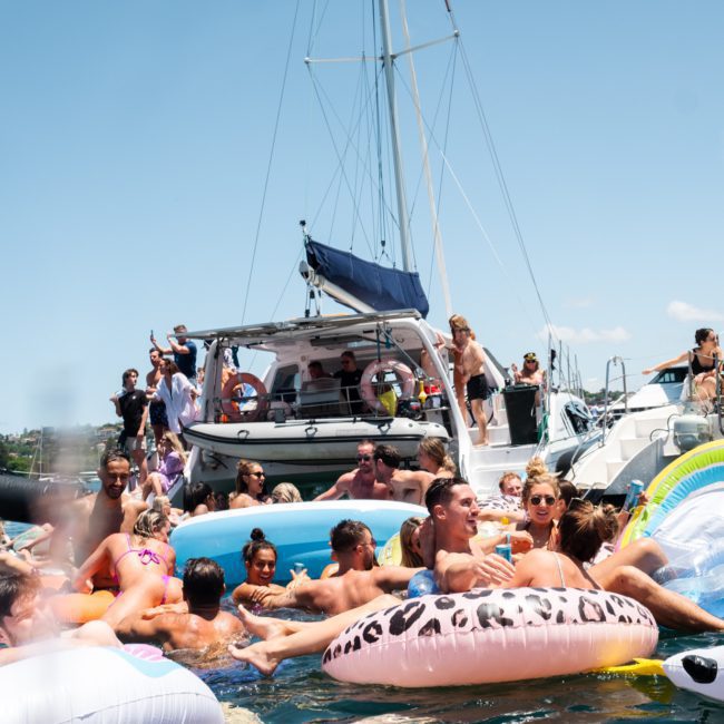 A group of people enjoying a sunny day on the water with various inflatable floats around multiple docked boats, part of a lively Sydney boat party hire.