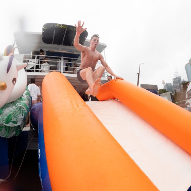 A person in swimwear slides down an orange and white inflatable slide from a luxury yacht hired in Sydney, with one hand raised mid-air.