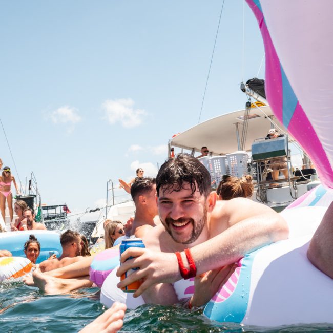 A man smiles and holds a can while floating on an inflatable in the water, surrounded by other people and boats at a lively catamaran party Sydney.
