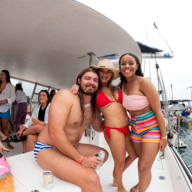 Three people pose together on a private yacht charter in Sydney Harbour, smiling at the camera. The man on the left is shirtless in striped swim trunks, the woman in the middle wears a red bikini and hat, and the woman on the right wears a colorful outfit.