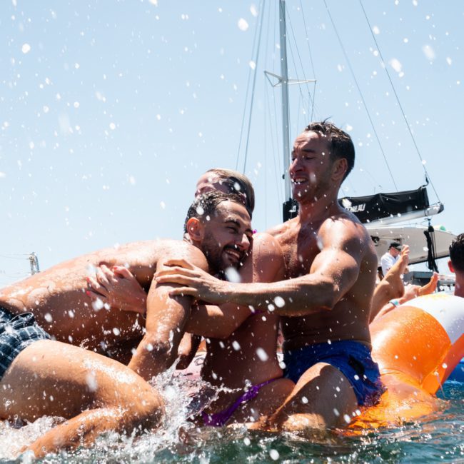 Three men in swimwear are playfully hugging in the water near boats, with water splashing around them and other people in the background enjoying the sunny day during a lively Sydney boat party hire.