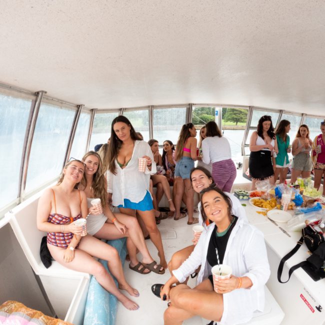 A group of people are enjoying a casual gathering on a private yacht charter in Sydney Harbour. Some are seated, holding drinks and smiling, while others are standing and chatting near a table with food and snacks.