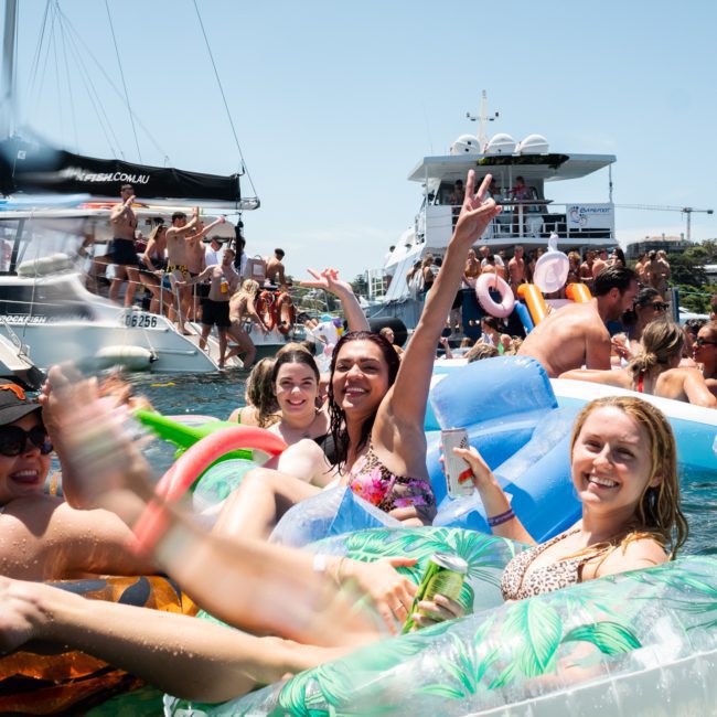 A group of people on inflatable floats enjoying a sunny day on the water near boats. Some people are raising their arms and smiling, making it the perfect scene for a Sydney boat party hire.