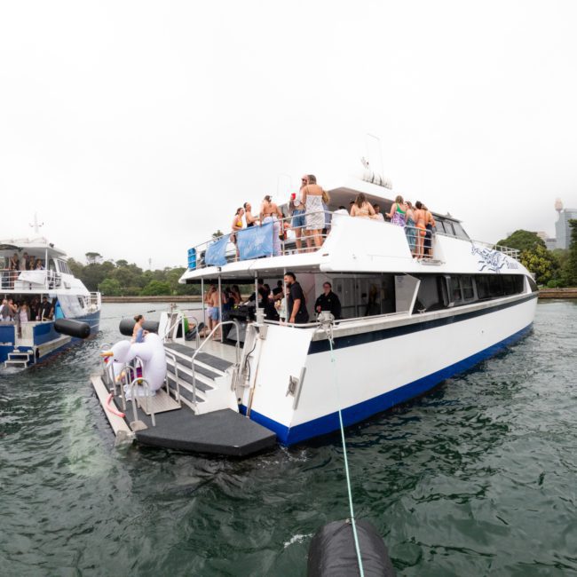 Two boats are anchored near a city shoreline. One boat, available for private yacht charter in Sydney Harbour, has a crowd of people on its deck enjoying a lively gathering. A smaller boat with a few individuals is nearby. The water is calm and the sky cloudy.