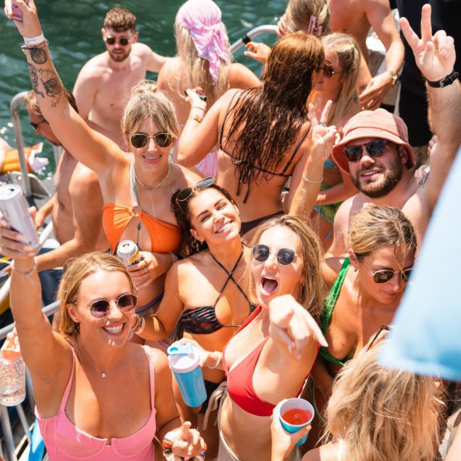 A group of people dressed in swimwear are gathered on a catamaran party Sydney, enjoying drinks and posing for the camera on a sunny day. Some are smiling, and others are raising their hands in celebration.
