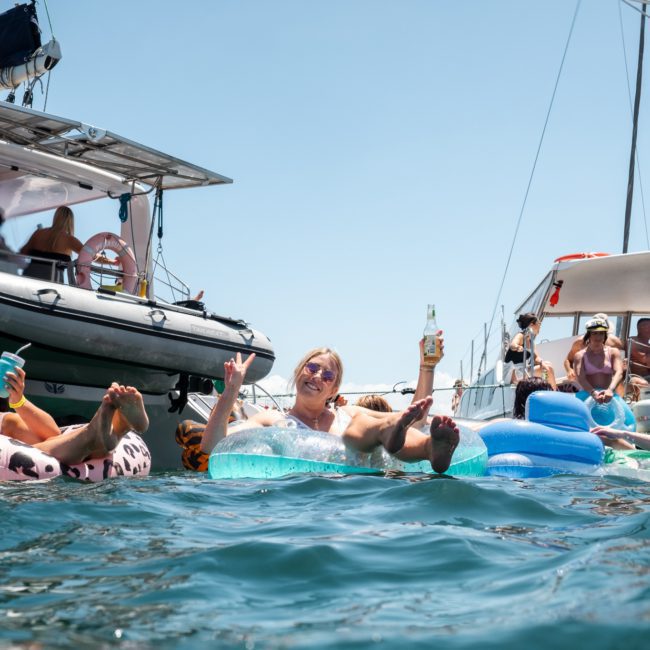 People float on inflatable rafts, holding drinks, near boats anchored in clear blue water under a sunny sky during a catamaran party Sydney event.