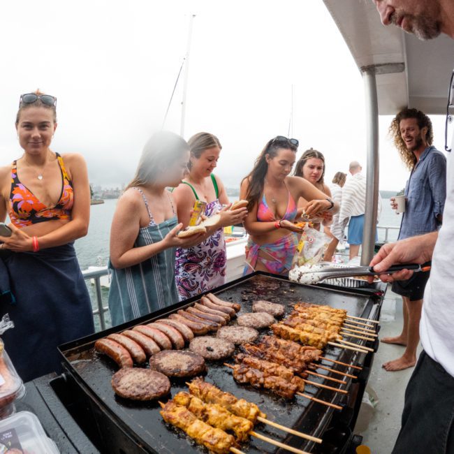 A group of people in swimwear gathers around a grill on a luxury yacht in Sydney Harbour. Various meats, including hamburgers, sausages, and skewers, are being cooked. One person is actively grilling the food at this lively Sydney boat party hire.