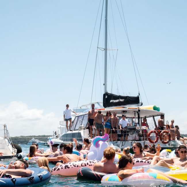 People are gathered on a private yacht charter in Sydney Harbour and in inflatable floaties on the water, enjoying a sunny day.