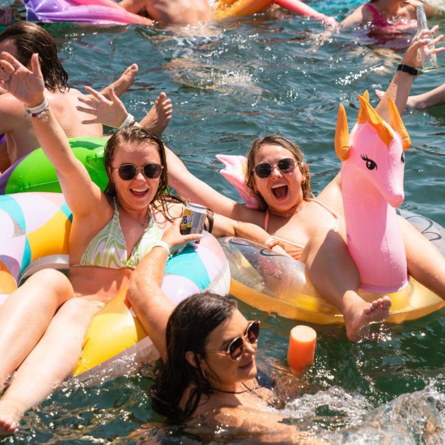People enjoying a sunny day in the water with inflatable pool floats, including a unicorn and a colorful ring, while smiling and raising their arms during a luxury yacht hire Sydney event.
