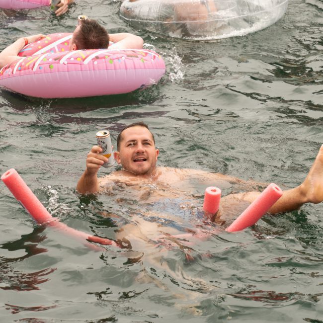 A man floating in water with pool noodles and holding a can, surrounded by other people on inflatable pool floats, reminiscent of a lively Sydney boat party hire.