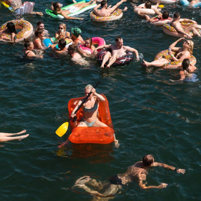 A group of people floats and swims in a body of water, many using various inflatable rafts and tubes. One person in the center paddles an orange inflatable raft with a yellow oar, enjoying what could be an ideal setting for a Sydney boat party hire.
