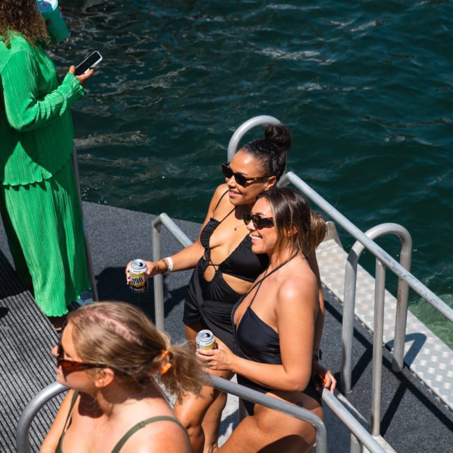 A group of people in swimwear enjoy a sunny day on a private yacht charter Sydney Harbour, with two women in black swimsuits holding drinks while standing near a railing by the water.