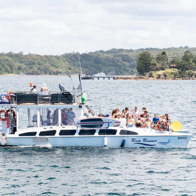 A white boat named "Rum Runner" with people on board is sailing on a body of water, framed by a small island and forested shore in the background, perfect for a luxurious yacht hire in Sydney.