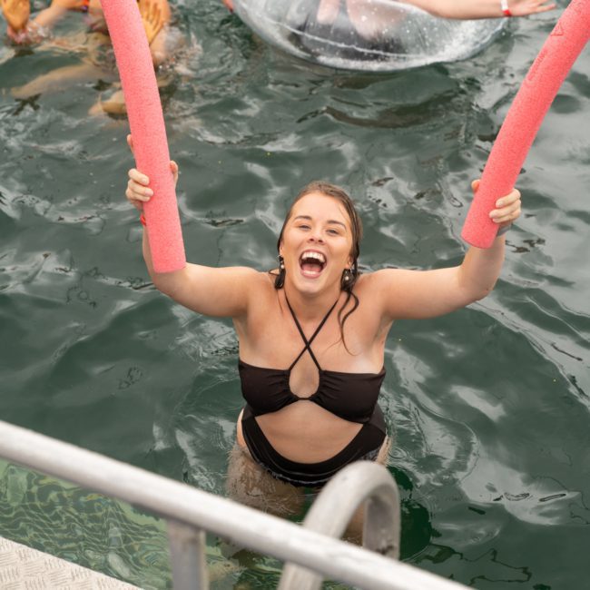 A smiling woman in a black swimsuit holds two pink pool noodles while standing in a body of water, with other people floating nearby. The scene evokes the carefree ambiance of a private yacht charter on Sydney Harbour.
