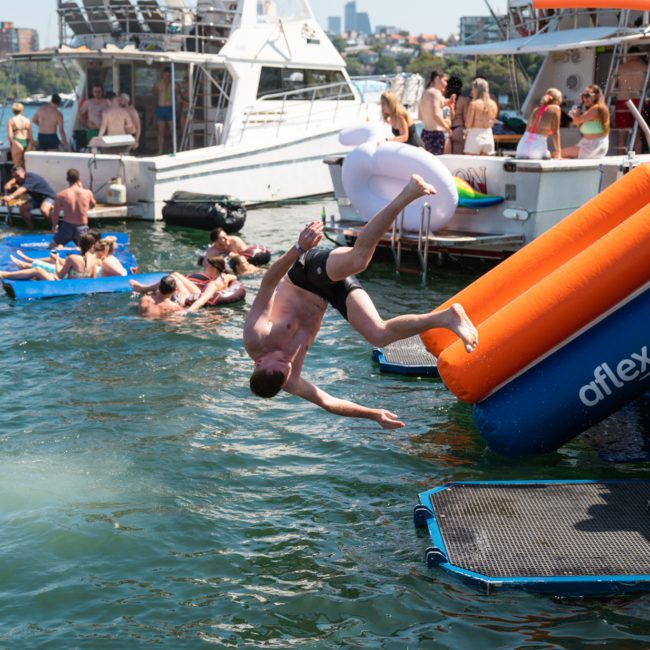 Person in mid-air after sliding down an inflatable orange and blue slide into a body of water, surrounded by people on boats and in the water during a corporate boat event in Sydney Harbour.