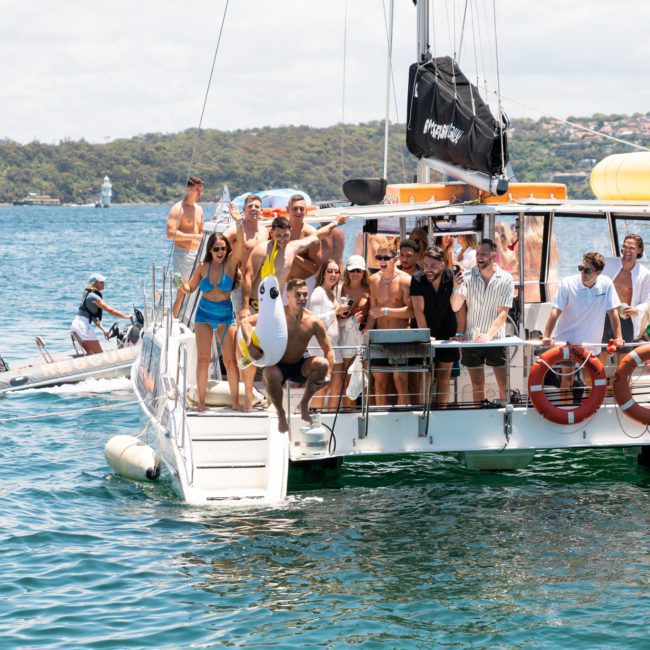 A group of people on a luxury yacht hire Sydney enjoy a sunny day on the water, waving and posing for the camera, with a small motorboat nearby and a hilly shoreline in the background.