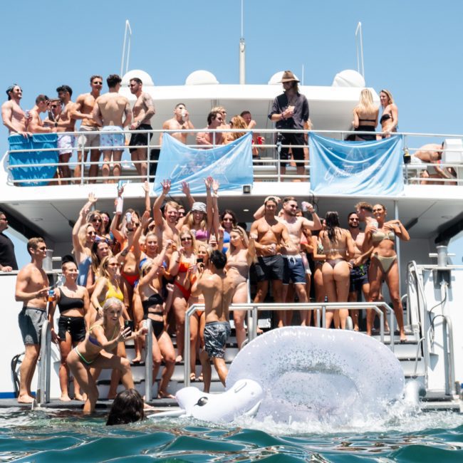 A large group of people in swimwear enjoying a sunny day on a catamaran party in Sydney, with some standing on the deck and others in the water near an inflatable swan.