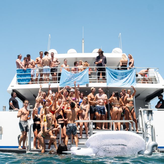 A large group of people in swimwear are gathered on the deck of a boat, some standing and some raising their hands in the air. The boat is on the water, and it's a sunny day. Perfect for corporate boat events Sydney or DJ boat hire Sydney.