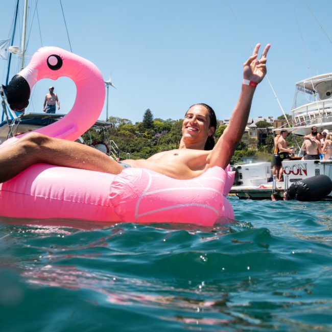A man relaxes on a pink flamingo float in the ocean, making a peace sign with his hand. In the background, groups of people are enjoying a catamaran party Sydney on boats and yachts.