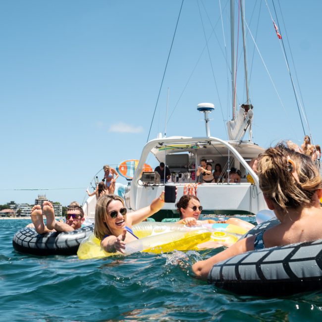A group of people relax on inflatable tubes in the water near a luxury yacht hire Sydney on a sunny day.