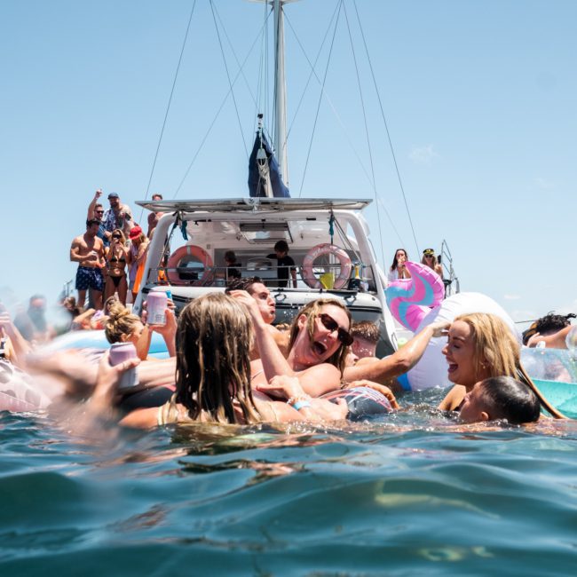 A group of people are enjoying themselves on and around inflatable floats in the water near a boat on a sunny day, with Sydney boat party hire providing the perfect backdrop for fun.