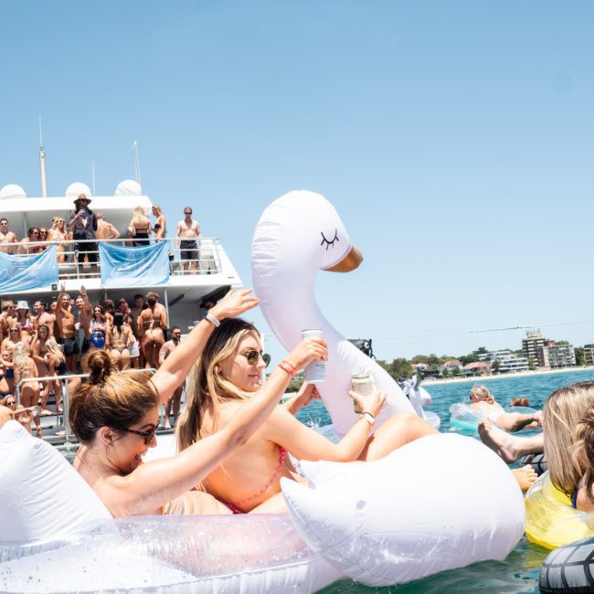 People in swimwear are at a crowded boat party in Sydney. Some are seen on large inflatable swans in the water, while others are on the boat. The sky is clear and sunny, making it a perfect day for a DJ boat hire Sydney experience.