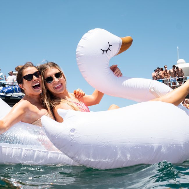 Two women sit on an inflatable swan float in the water, smiling and holding drinks, with a crowd of people enjoying a Sydney boat party hire on a nearby boat in the background.