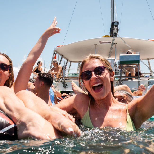 A group of people in swimwear enjoying themselves in the water near a boat, with some raising their arms and smiling during a Corporate boat event in Sydney.