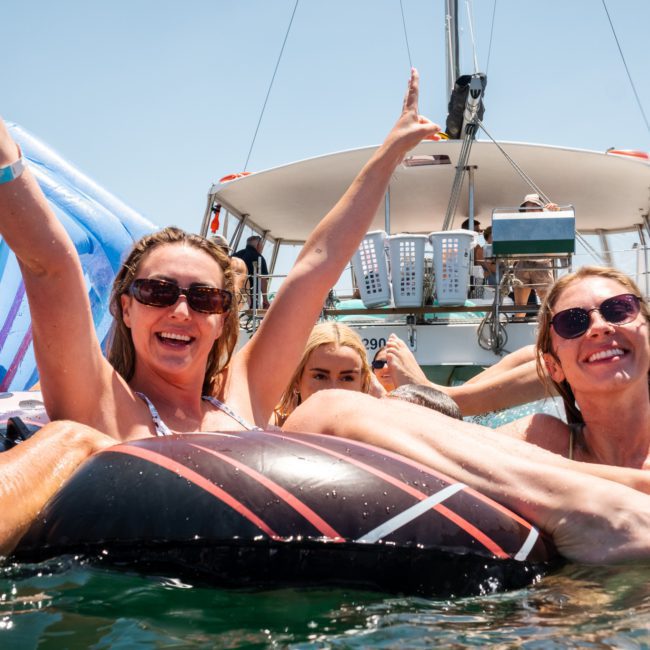 A group of people are enjoying themselves on inflatable floats in the water near a boat. Two women in the foreground are smiling, raising their arms, and holding drinks at a lively catamaran party in Sydney.