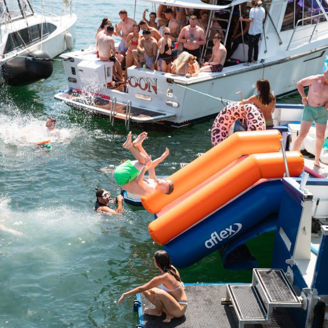 People enjoying a lakeside party with boats, swimming, and a water slide. A person is mid-air after sliding down an inflatable slide while others swim or relax on the boats. The atmosphere feels like a Catamaran party in Sydney with friends reveling in the fun and luxury of the event.