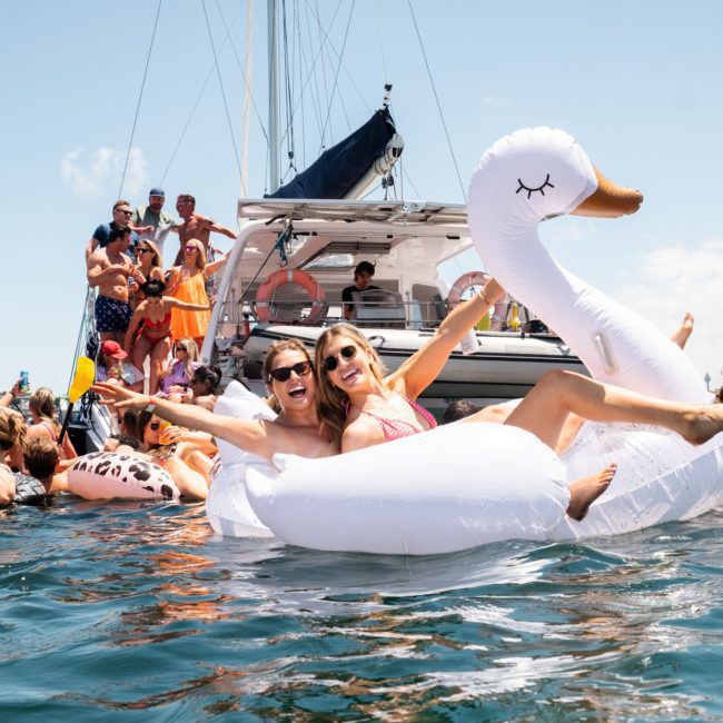 Two people smile and pose in a large swan pool float surrounded by others in the water near a docked sailboat on a sunny day, perfect for a Luxury yacht hire Sydney experience.