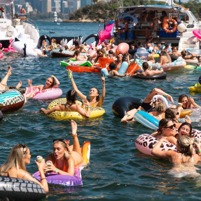A large group of people enjoying a sunny day on the water with a variety of pool floats, near boats, and a scenic city skyline in the background. They are partaking in a luxury yacht hire Sydney experience, making the day truly special with picturesque views and elegant surroundings.
