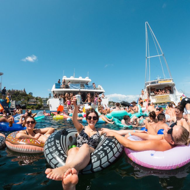 People relaxing on colorful inflatable floats in the water while boats are anchored nearby on a sunny day during a luxury yacht hire Sydney event.