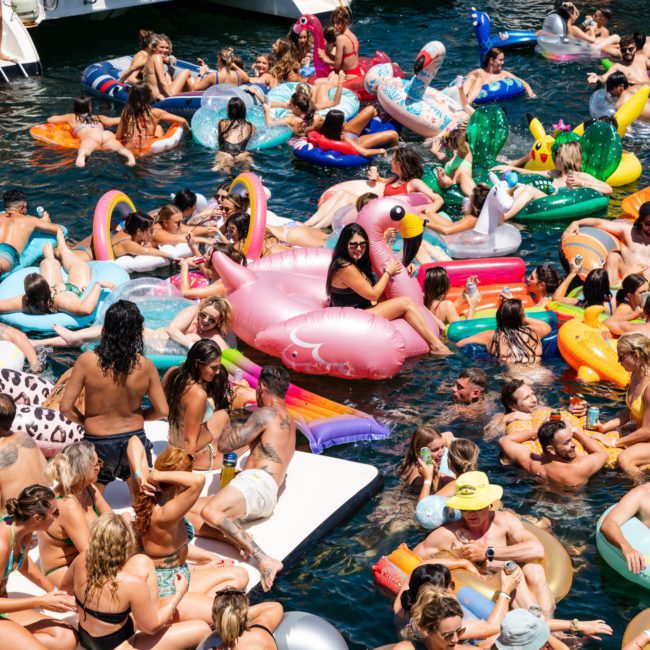 A lively outdoor party on the water with people on various inflatable floats, including a large pink flamingo, surrounded by boats and enjoying a sunny day. Perfect for those looking to experience the fun of a Catamaran party in Sydney.