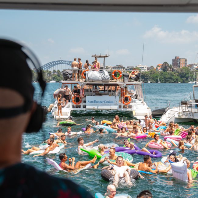 A group of people enjoying a sunny day on a lake with many floating on inflatables, surrounded by boats, with a bridge in the background. Perfect for a private yacht charter Sydney Harbour or even a Sydney boat party hire.