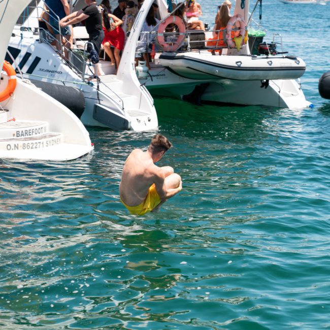 A person in yellow shorts is mid-air, curled in a ball, about to splash into the water from a boat. Other boats with people are in the background on a sunny day, enjoying what looks like a fun corporate boat event in Sydney.