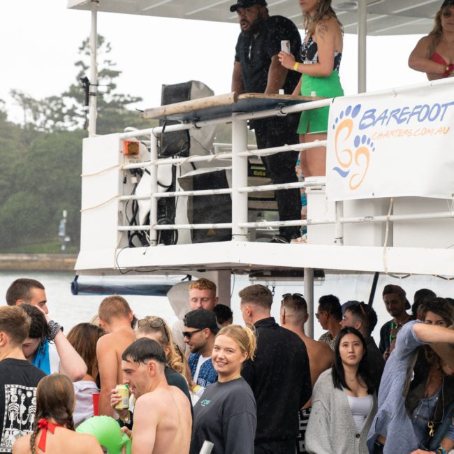People gathered on a boat, some in swimsuits, socializing. One woman in a green skirt stands near the railing on the upper deck. The luxury yacht hire Sydney has a "Barefoot" sign and appears to be at a party.