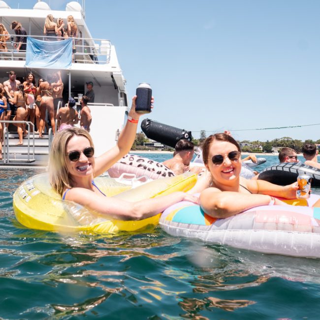 Two women relax on inflatable floats in the water, holding drinks. A Sydney boat party hire is visible on a nearby boat with many people gathered on the deck. The weather appears sunny and clear.