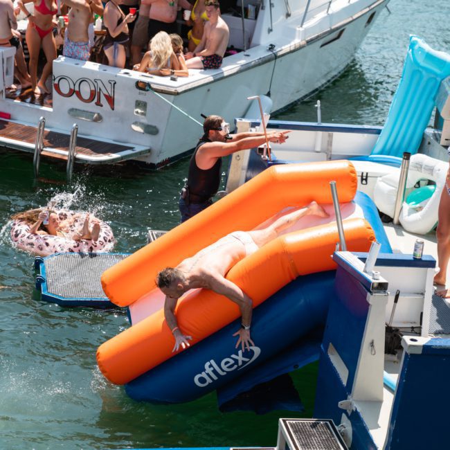 People enjoying water activities around boats, with some swimming and a man sliding down an inflatable slide into the water. Others are on board, standing or sitting on the boats during their private yacht charter Sydney Harbour.