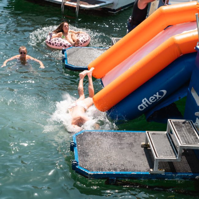 Person sliding down an inflatable slide into a body of water, where two other individuals are floating nearby on pool floats, all while enjoying a DJ boat hire Sydney.