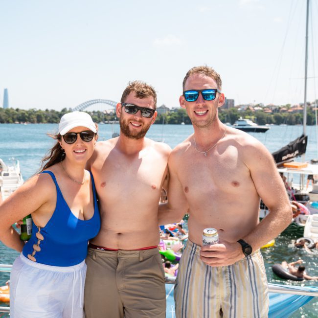 Three people in swimwear stand on a private yacht charter in Sydney Harbour with water, other boats, and a bridge in the background. They are smiling and wearing sunglasses on a sunny day.