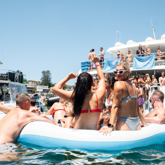 People on an inflatable raft and boat enjoying a sunny day on the water. There are other boats and individuals in the background, including a catamaran party Sydney.