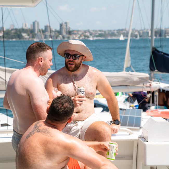 Three men relax on a private yacht charter under a sunny sky, engaging in conversation while holding drinks. The backdrop features a scenic view of the water and distant cityscape.