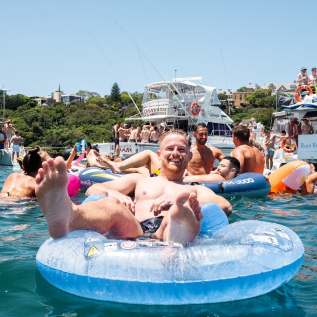A person floats on a blue inflatable ring in the water, surrounded by several other people and boats. The weather is sunny, and they appear to be enjoying a day out on the water, perhaps after renting a luxury yacht hire Sydney for some added fun.