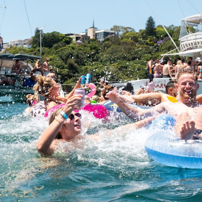 People enjoying a sunny day on the water, floating on inflatable tubes and splashing around near boats anchored close to the shore, with a hint of excitement in the air from those who opted for a DJ boat hire Sydney.