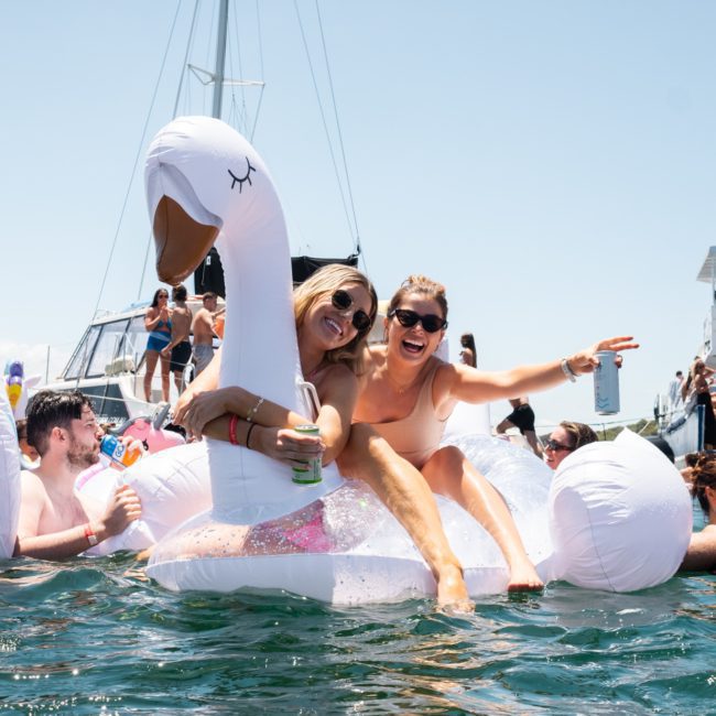 Two women smile and pose on a large inflatable swan float in the water, surrounded by people enjoying a sunny day with boats in the background, epitomizing a perfect day made even better with Sydney boat party hire.