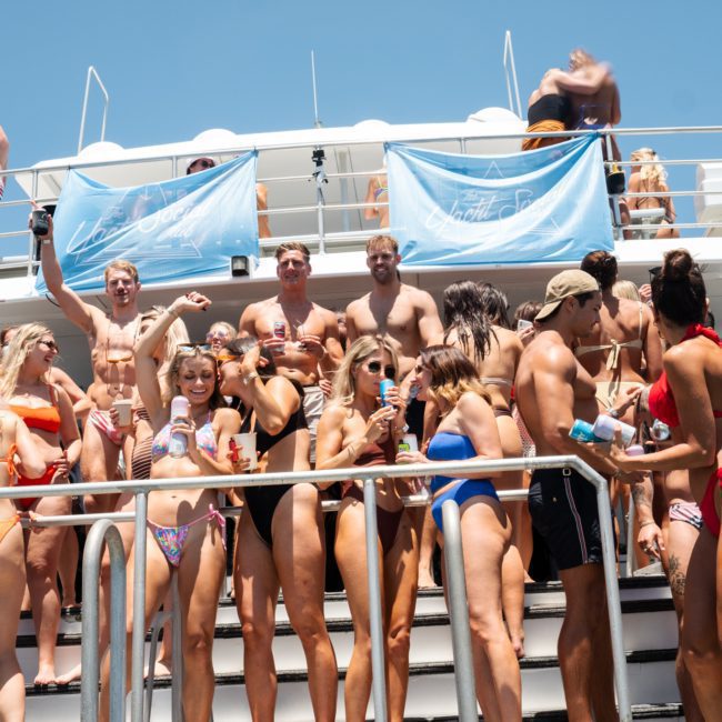 A group of people in swimwear gathered on a luxury yacht hire in Sydney, some holding drinks and posing for the camera. The boat has banners with text and a clear blue sky in the background.