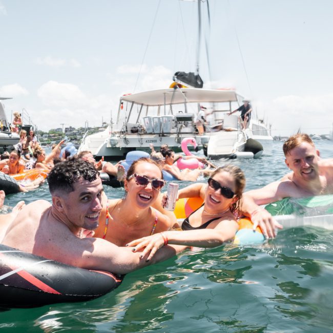 A group of people swim and relax on inflatable floats near anchored boats on a sunny day, enjoying the perfect setting for a Catamaran party in Sydney.