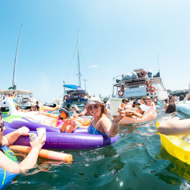 People relax in a sunny bay on colorful inflatable floats, holding drinks, with boats anchored nearby, enjoying a lively catamaran party in Sydney.